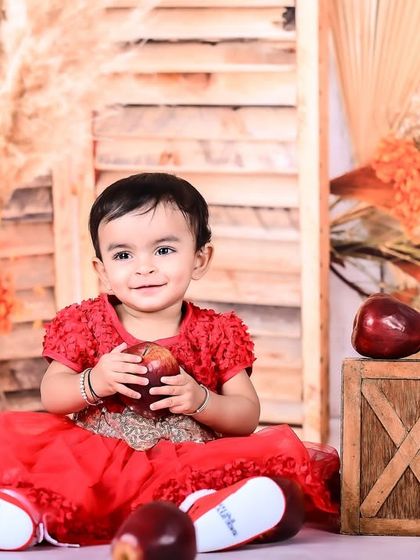 A baby girl in a red dress sitting in a rustic apple-themed setup.