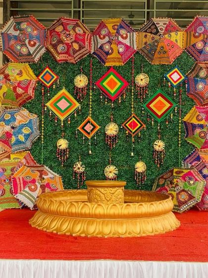 An elaborate and colorful backdrop for a Haldi ceremony, featuring a green wall decorated with Rajasthani umbrellas and a traditional 'urli' for the rituals.