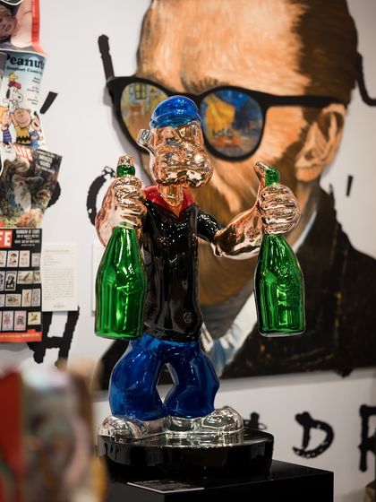 This sculpture of Popeye, rendered in metallic tones and holding green bottles, reimagines the classic cartoon character as a modern icon. It stands before a contemporary portrait, creating a layered visual effect.
