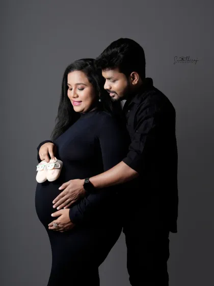 A sweet couple portrait in matching black outfits. The husband gently holds his wife's bump as they hold up a pair of tiny baby shoes, symbolizing their shared excitement.