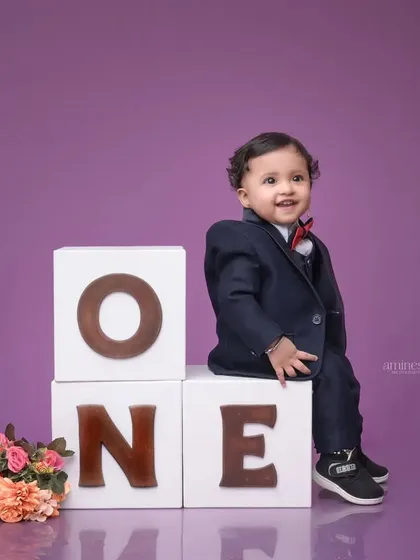 Celebrating a first birthday milestone with a classic studio portrait. A smartly dressed one-year-old boy sits proudly on large "ONE" blocks against a clean purple background, creating a timeless memory of this special day.