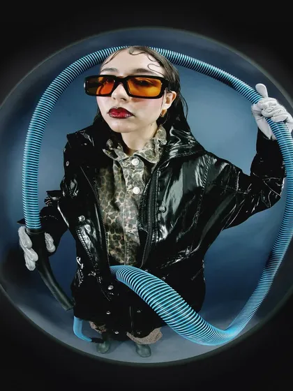 A similar setup with the blue tube, but with a different pose and expression. The combination of the shiny black jacket, animal print shirt, and orange glasses creates a bold, eclectic look.