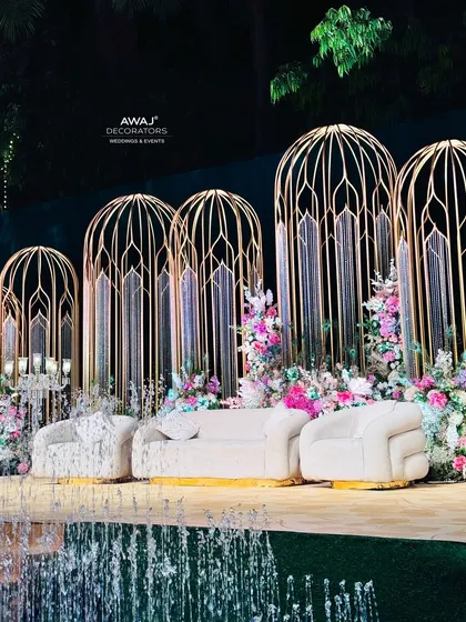A stunning stage setup featuring ornate golden cage-like structures, pastel floral arrangements, and a cascading water feature in the foreground.