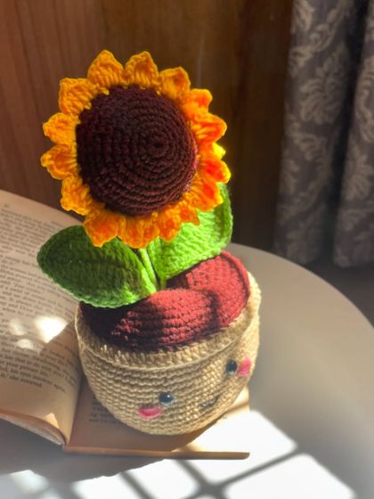 A close-up of my smiling sunflower pot, sitting next to an open book. It's the perfect little desk buddy to keep you company while you work or study.