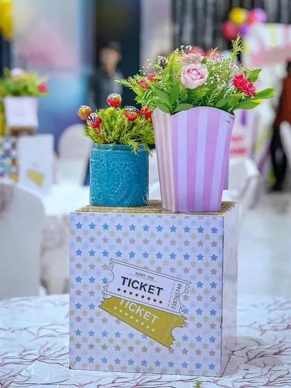 A close-up of a carnival table centerpiece. It features a star-patterned box with a ticket graphic, topped with fresh flowers in a striped container and a jar of lollipops, showing my attention to every small detail.