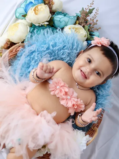 This happy baby girl lies in a basket filled with soft fabrics and flowers, smiling and reaching for the camera.