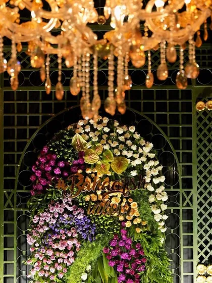A close-up of a floral arrangement with a custom 'Bhavna & Bhupendra' sign, set against a green lattice backdrop and lit by a chandelier.