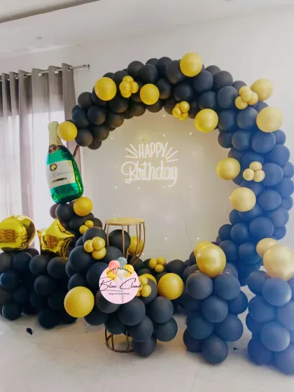 A classic and stylish black and gold balloon ring backdrop perfect for milestone birthdays or sophisticated parties. This setup includes a 'Happy Birthday' neon sign and a champagne bottle foil balloon.