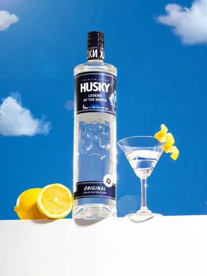 A bright and refreshing shot of a vodka bottle against a sky-blue background with fluffy clouds. The composition with a classic martini glass and fresh lemon evokes a feeling of a perfect, sunny day.