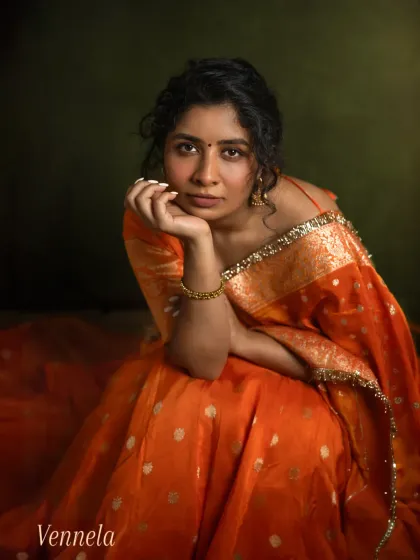 This close-up portrait of Vennela in her vibrant orange saree focuses on her direct and engaging gaze. The rich colors and soft lighting create a warm, inviting image that captures her confidence and personality.
