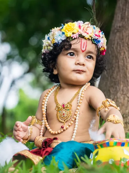 A baby boy dressed as Krishna sits in the grass, curiously playing with a pot of 'butter' (cotton).