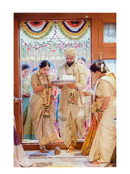 The couple performs a traditional ceremony at the doorway. This image captures the groom's full attire, including the dhoti and shawl, in a moment of cultural significance.