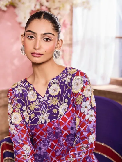 A close-up portrait in a colorful purple and red embroidered top. The makeup is clean and fresh, with a soft pink blush and defined brows to frame the face.