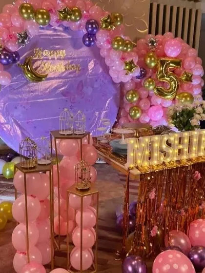 A glamorous pink and purple 5th birthday party for Mishika. The room is filled with metallic balloons, a large backdrop, multiple pedestals for treats, and light-up letters for the name.