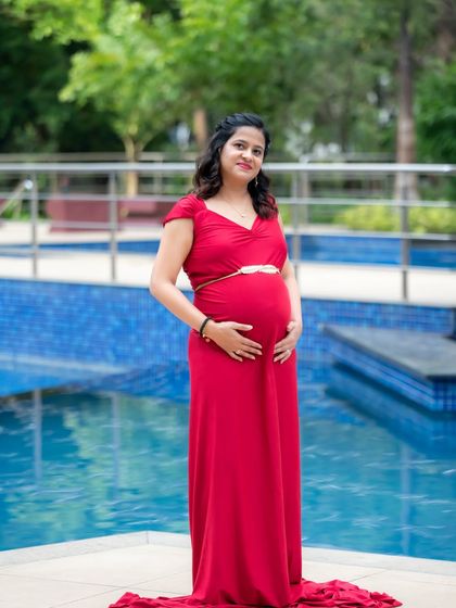 A serene solo portrait of a mom-to-be in a classic red dress, standing by a bright blue swimming pool. It's a simple yet elegant setting for a maternity shoot.