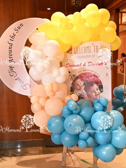 A celestial-themed welcome sign for twins Devansh and Devdutt's "First Trip Around the Sun." The sign features a crescent moon, a sun, and a photo of the birthday boys, framed by blue, yellow, and peach balloons.