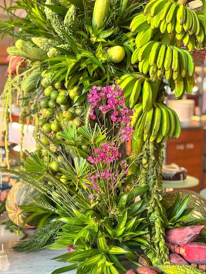 A close-up of a nature-inspired dining display. Here, green bananas, limes, and other vegetables are artfully combined with pink flowers and diverse foliage to elevate the dining experience.