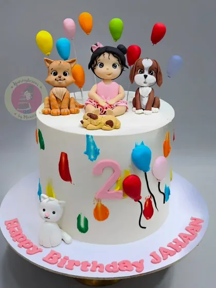 A colorful and happy cake for Jahaan's 2nd birthday, featuring a figurine of her with her pet cat and dog, surrounded by balloons.