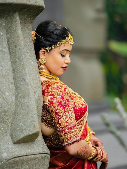 A bride leans against a stone pillar, her red saree creating a beautiful contrast. The intricate embroidery on her blouse is a testament to our craftsmanship.