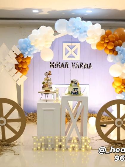 A charming farm-themed birthday party with a barn-shaped backdrop and a color scheme of light blue, white, and brown. The decor includes cutouts of a cow and sheep, large wagon wheels, and illuminated letters on a bed of hay.