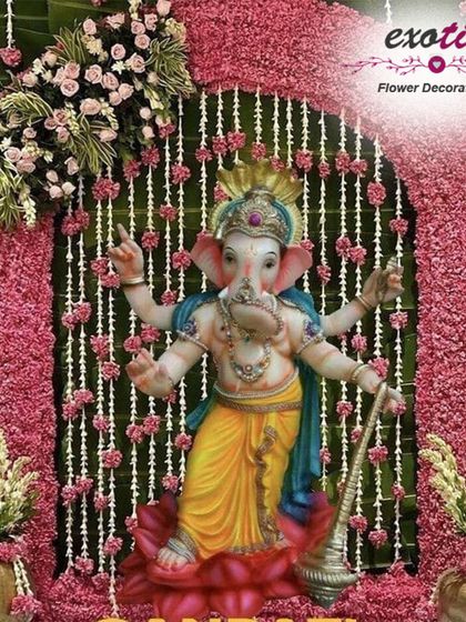 A vibrant and traditional Ganpati decoration featuring a stunning idol against a backdrop of pink flowers and hanging pearl strings. The dense floral arch and side arrangements create a rich and festive look, perfect for welcoming Bappa into your home.