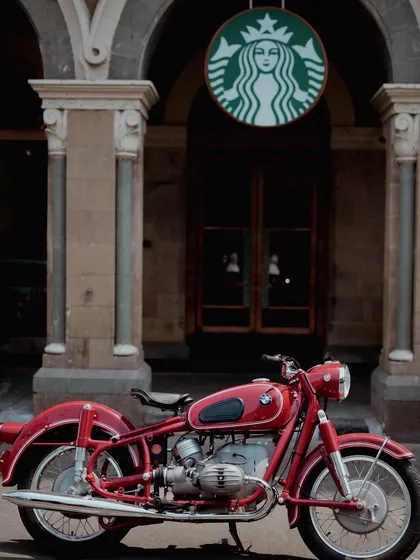 A striking vintage red BMW motorcycle is parked outside a modern Starbucks, creating a beautiful contrast between classic German engineering and contemporary culture. This urban automotive shot highlights the bike's timeless appeal in a familiar city setting.