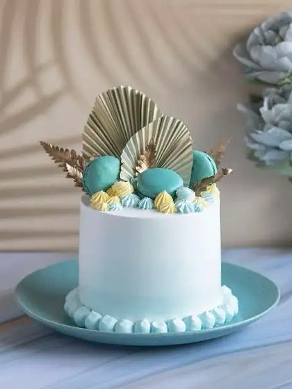 An elegant and modern birthday cake design. This smooth, light blue cake is topped with macarons and decorative dried palm leaves for a chic, contemporary look.