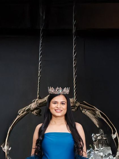 A close-up portrait of a smiling mother-to-be wearing a crown, seated on an ornate swing. The dark background ensures all attention is on her happy expression.
