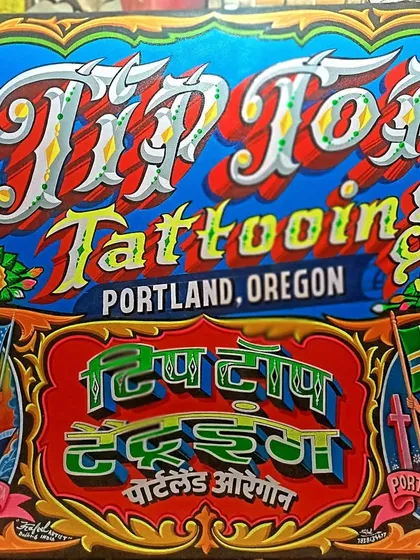A sign for Tip Top Tattooing in Portland, Oregon, painted in both English and Hindi. The artwork includes American and local flags, showing a blend of cultures.