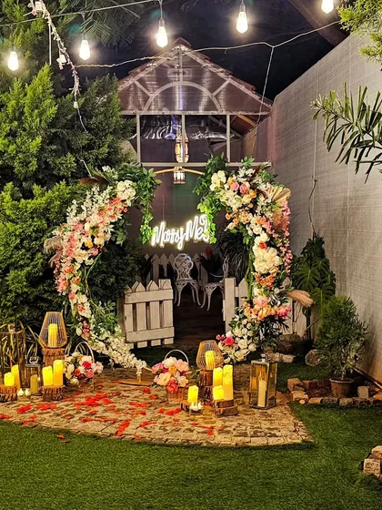 This cozy and intimate garden nook is transformed for a proposal. A circular floral arch frames a small seating area, surrounded by lush greenery and illuminated by candles and a "Marry Me" sign.