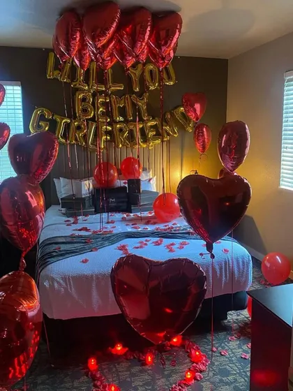 A bold and romantic proposal setup in a hotel room. The room is filled with red heart-shaped foil balloons and rose petals, with a "Will You Be My Girlfriend" foil balloon message above the bed.