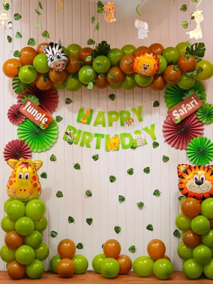 A classic jungle safari birthday theme. The backdrop is decorated with green paper fans and leaf cutouts, framed by balloon pillars with giraffe and tiger foil balloon toppers.