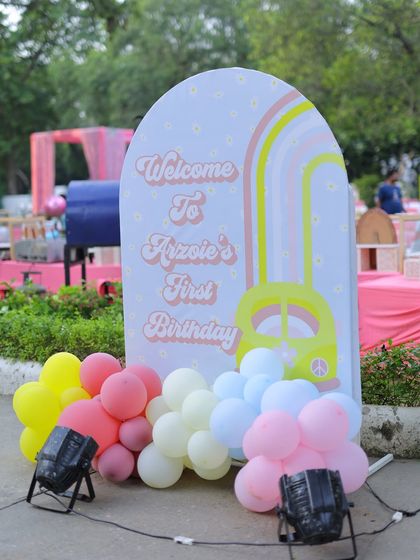 A groovy "Welcome to Arzoie's First Birthday" sign. The retro font, peace-sign bus graphic, and pastel rainbow perfectly match the sunshine theme and are framed by colorful balloons.