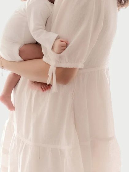 A close-up detail shot of a mother in a white dress holding her baby. The focus on the tiny hands and the way she holds her child speaks of the protective and nurturing nature of a mother's love.