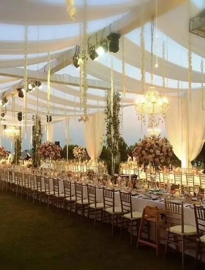 An exquisite long-table dining setup for an outdoor wedding reception. The tables are arranged under a canopy of white drapes and hanging floral strings, with chandeliers adding a touch of elegance for an evening under the stars.