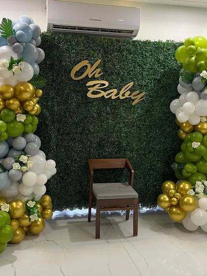 An elegant "Oh Baby" shower setup with a lush green foliage wall. The backdrop is framed by beautiful balloon columns in shades of green, gold, grey, and white, creating a fresh and natural look.