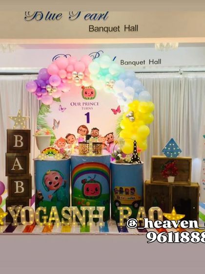A colorful Cocomelon-themed first birthday party. The decor features a large JJ cutout, a rainbow balloon arch, and plinths decorated with characters and scenes from the popular show.