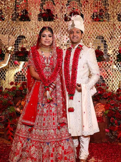 A portrait of the couple in their wedding attire. The backdrop is a rich tapestry of red flowers and intricate white jaali work, creating a grand and traditional wedding stage.