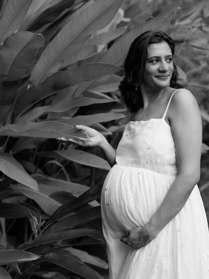 A beautiful black and white portrait of an expecting mother in a simple white dress, standing amongst large green leaves. It's a serene and natural shot.