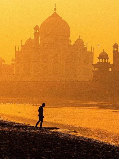 A lone figure walks along the riverbank with the Taj Mahal shrouded in a golden, hazy sunrise. The image is atmospheric and evokes a sense of solitude and grandeur.