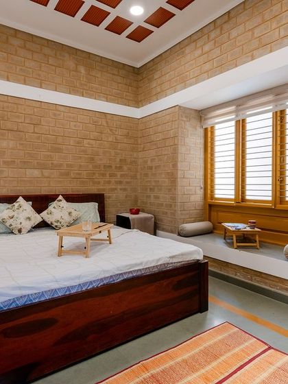 A bedroom in Audumbara, featuring exposed CSEB walls and a filler slab ceiling with clay tiles. A cozy bay window seat provides a perfect reading nook, lit by natural light.