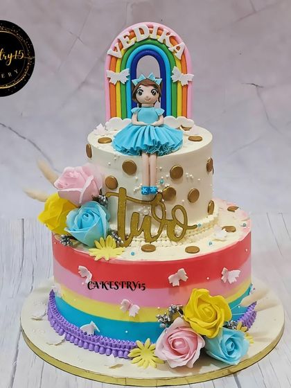 A beautiful two-tier doll-themed cake for a second birthday. This vanilla cake features a rainbow-striped bottom tier, a doll topper, and is adorned with colorful faux roses.
