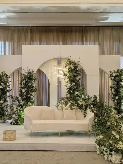 A minimalist and elegant stage design for a modern reception or Nikkah ceremony. I used clean white panels with archways, accented with fresh greenery and soft lighting to create a sophisticated and serene ambiance.
