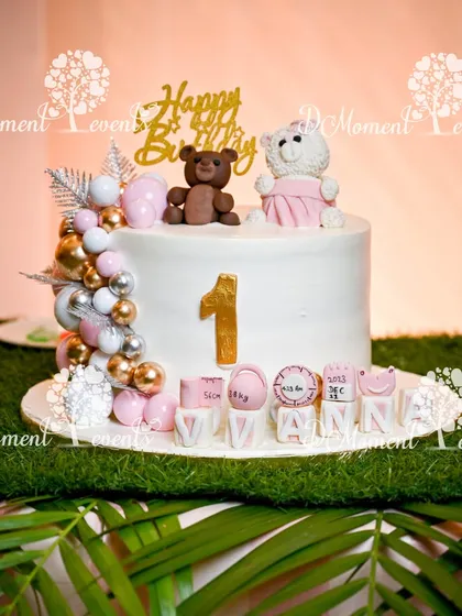 A close-up of a teddy bear-themed first birthday cake. The cake is topped with two cute teddy bears and sits on a bed of artificial grass.