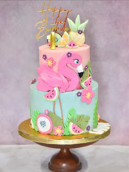 A tropical two-tier cake for a flamingo-themed first birthday. This vibrant creation is adorned with a hand-sculpted pink flamingo, fondant pineapples, watermelons, and tropical leaves for a fun, summery celebration.