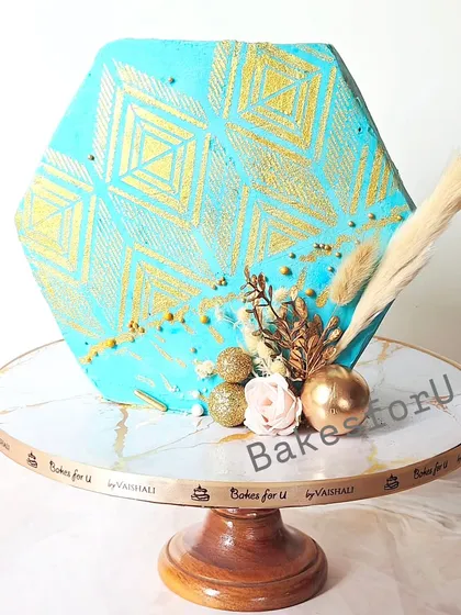 A unique hexagonal cake with a striking blue and gold geometric stencil pattern. The cake is finished with dried grasses and gold spheres for a modern, boho-chic look.