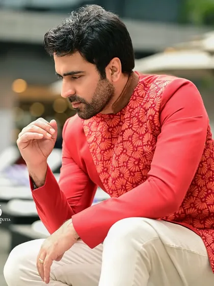 A thoughtful, candid-style portrait of Abir Chatterjee in a vibrant red jacket. The outdoor, poolside setting provides a relaxed backdrop for this contemporary ethnic menswear look.