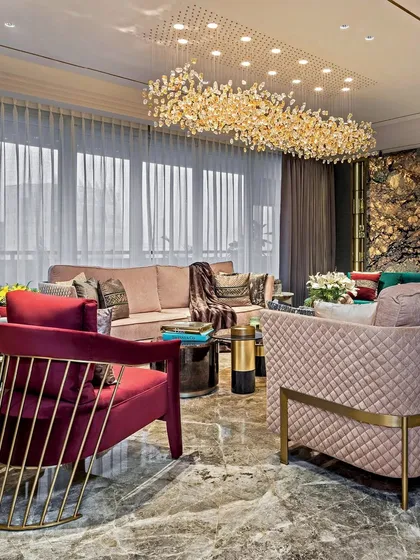 A luxurious living room featuring a mix of seating, including a quilted armchair and a plush sofa with red accents. The space is illuminated by a large, intricate chandelier, creating a warm and glamorous atmosphere.