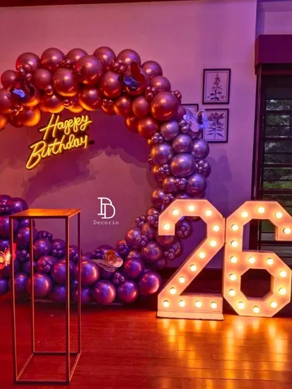 A vibrant 26th birthday setup with a moody and stylish color palette. The decor features a circular arch of metallic rose gold and purple balloons, complemented by large '26' light-up numbers and a central neon sign.