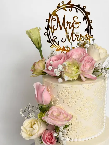 A close-up of a floral dream wedding cake. This shot highlights the delicate texture on the top tier and the beautiful arrangement of soft pink and white roses, topped with a "Mr & Mrs" charm.